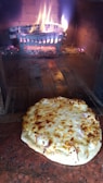 A freshly baked pizza with melted cheese sits on the floor of a brick oven. Behind it, there is a glowing fire with burning logs held in a metal grate, emitting a warm and inviting atmosphere.