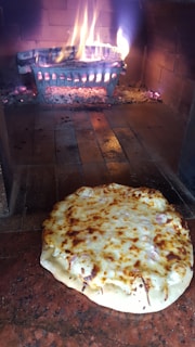 A freshly baked pizza with melted cheese sits on the floor of a brick oven. Behind it, there is a glowing fire with burning logs held in a metal grate, emitting a warm and inviting atmosphere.