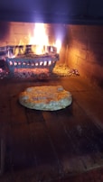 A wood-fired oven interior with a pizza being baked on the brick floor. The flames and embers provide an authentic, rustic cooking environment. The pizza appears to be a simple cheese variety, partially melted and starting to bubble.