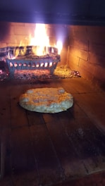 A wood-fired oven interior with a pizza being baked on the brick floor. The flames and embers provide an authentic, rustic cooking environment. The pizza appears to be a simple cheese variety, partially melted and starting to bubble.