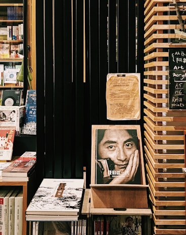 A cozy bookstore corner featuring shelves filled with various books. Prominent is an expressive book cover displaying a black-and-white photo of a man resting his face on his hand. The setting includes wooden shelves and dark slats, creating a warm and contemplative atmosphere. A small chalkboard lists items like art supplies and refill prints.