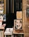 A cozy bookstore corner featuring shelves filled with various books. Prominent is an expressive book cover displaying a black-and-white photo of a man resting his face on his hand. The setting includes wooden shelves and dark slats, creating a warm and contemplative atmosphere. A small chalkboard lists items like art supplies and refill prints.
