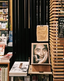A cozy bookstore corner featuring shelves filled with various books. Prominent is an expressive book cover displaying a black-and-white photo of a man resting his face on his hand. The setting includes wooden shelves and dark slats, creating a warm and contemplative atmosphere. A small chalkboard lists items like art supplies and refill prints.