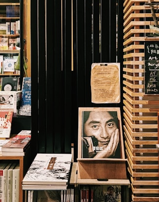 A cozy bookstore corner featuring shelves filled with various books. Prominent is an expressive book cover displaying a black-and-white photo of a man resting his face on his hand. The setting includes wooden shelves and dark slats, creating a warm and contemplative atmosphere. A small chalkboard lists items like art supplies and refill prints.