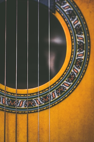 A close-up view of a guitar's sound hole, surrounded by intricate and colorful inlays. The metallic strings run across the image vertically, creating a sharp contrast against the warm tones of the wood and decorative pattern.