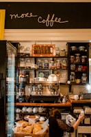 A cozy coffee shop with jars of different teas and coffee beans lining the shelves. The central focus is a large espresso machine surrounded by cups and other coffee-making equipment. Below, there is a counter with a display of pastries. A person is seen reaching into a small fridge beneath the counter.