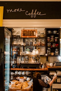 A cozy coffee shop with jars of different teas and coffee beans lining the shelves. The central focus is a large espresso machine surrounded by cups and other coffee-making equipment. Below, there is a counter with a display of pastries. A person is seen reaching into a small fridge beneath the counter.