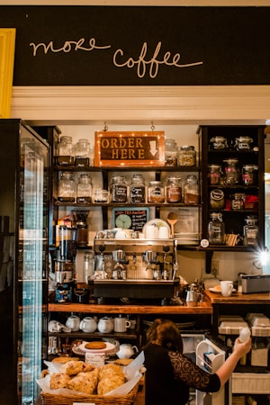A cozy coffee shop with jars of different teas and coffee beans lining the shelves. The central focus is a large espresso machine surrounded by cups and other coffee-making equipment. Below, there is a counter with a display of pastries. A person is seen reaching into a small fridge beneath the counter.