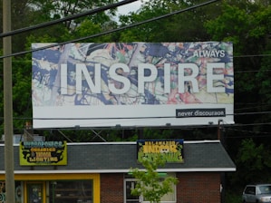 A large billboard with an abstract background and the word 'INSPIRE' prominently displayed in white letters. The words 'ALWAYS' and 'never discourage' accompany the main text. Below the billboard, a small store is visible, with a roof sign that reads 'Kalamazoo Indoor Gardening' and 'Hydroponics, Organics, Indoor Lighting' on a yellow background. The area is surrounded by greenery and trees.