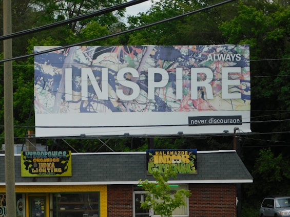 A large billboard with an abstract background and the word 'INSPIRE' prominently displayed in white letters. The words 'ALWAYS' and 'never discourage' accompany the main text. Below the billboard, a small store is visible, with a roof sign that reads 'Kalamazoo Indoor Gardening' and 'Hydroponics, Organics, Indoor Lighting' on a yellow background. The area is surrounded by greenery and trees.
