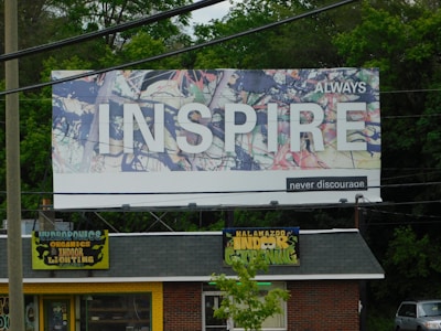 A large billboard with an abstract background and the word 'INSPIRE' prominently displayed in white letters. The words 'ALWAYS' and 'never discourage' accompany the main text. Below the billboard, a small store is visible, with a roof sign that reads 'Kalamazoo Indoor Gardening' and 'Hydroponics, Organics, Indoor Lighting' on a yellow background. The area is surrounded by greenery and trees.
