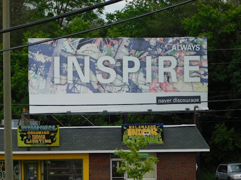 A large billboard with an abstract background and the word 'INSPIRE' prominently displayed in white letters. The words 'ALWAYS' and 'never discourage' accompany the main text. Below the billboard, a small store is visible, with a roof sign that reads 'Kalamazoo Indoor Gardening' and 'Hydroponics, Organics, Indoor Lighting' on a yellow background. The area is surrounded by greenery and trees.