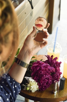 A person is holding a small mirror, reflecting their lips with red lipstick. On the table beneath, there is a bouquet of vibrant purple and white flowers. Nearby are two cold drinks in plastic cups with straws and a couple of tubes of lipstick. The setting seems to be outdoors with wooden paneling in the background.