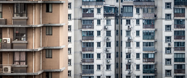 A close-up view of two adjacent apartment buildings, one with a beige facade and the other with a light gray exterior. Both buildings feature multiple balconies and numerous windows, with air conditioning units visible on some balconies. The architecture is modern and utilitarian.
