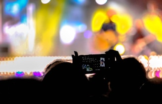 The image captures a crowd at a concert with vibrant, colorful lighting. The foreground shows the silhouette of a person holding up a smartphone to record or take photos of the performance. Blurred figures and bright stage lights create a lively and dynamic atmosphere.