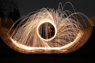 A dramatic display of steel wool photography inside a ramp, with sparks radiating outward in circular patterns. The background shows a dark night setting with silhouetted trees and yellow railings in the backdrop.