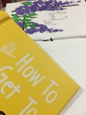 Close-up of a book cover in yellow with the text 'How To Get To'. In the background, there is a partial view of a floral illustration with purple flowers and green leaves.