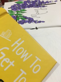 Close-up of a book cover in yellow with the text 'How To Get To'. In the background, there is a partial view of a floral illustration with purple flowers and green leaves.