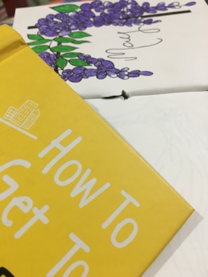 Close-up of a book cover in yellow with the text 'How To Get To'. In the background, there is a partial view of a floral illustration with purple flowers and green leaves.