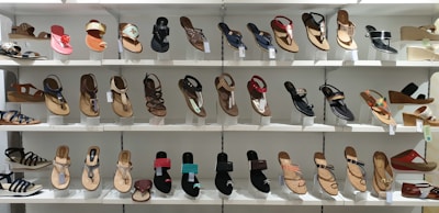 A display of various women's sandals arranged neatly on white shelves. The sandals are diverse in style, including flip-flops, strappy sandals, and wedges, showcasing a range of designs from casual to more decorative options.
