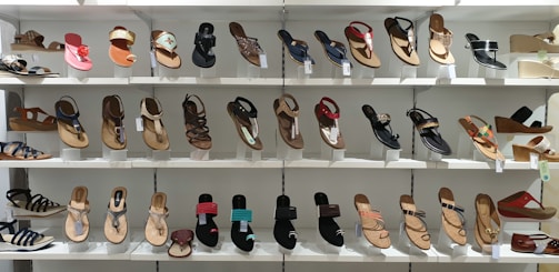 A display of various women's sandals arranged neatly on white shelves. The sandals are diverse in style, including flip-flops, strappy sandals, and wedges, showcasing a range of designs from casual to more decorative options.