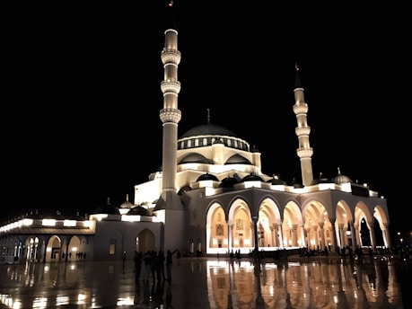 A beautifully illuminated mosque with multiple minarets stands prominently against a dark night sky. The structure features grand arches and domes, casting reflections on the polished surface of the courtyard, where people are gathered.