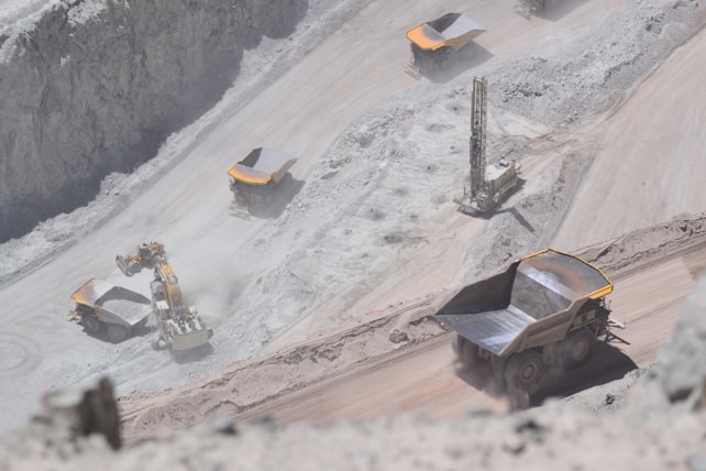 Several large dump trucks and an excavator are operating in an open-pit mine, with dusty and rocky terrain surrounding them. A drilling machine is also visible, indicating active mining activities.