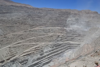 A large open-pit mine with terraced levels carved into a hillside, creating a massive and layered appearance. The landscape is mostly barren with dusty roads leading to various levels of the pit. The scene is devoid of vegetation, emphasizing the stark industrial activity.