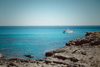 A coastal scene with clear blue water, rugged rocky shore, and a small boat with passengers. The sky is clear and the boat is relatively close to the shore, possibly touring or fishing.