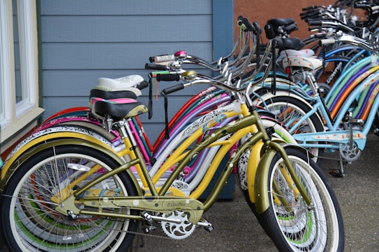 Several colorful cruiser bicycles are lined up against a building with blue siding. The bicycles have various patterns and designs on their frames, with a prominent green and yellow bike in the foreground. The concrete ground adds a neutral base to the vibrant colors of the bicycles.