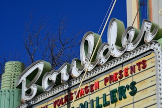 A retro-style marquee sign with the word 'Boulder' in large cursive font, surrounded by LED bulbs. A blue sky and leafless tree branches are visible in the background. The sign also includes words in red and green letters, such as 'VOLKSWAGEN PRESENTS' and 'MILLER'S'. The structure has an Art Deco design, featuring pastel colors like mint green.
