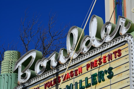 A retro-style marquee sign with the word 'Boulder' in large cursive font, surrounded by LED bulbs. A blue sky and leafless tree branches are visible in the background. The sign also includes words in red and green letters, such as 'VOLKSWAGEN PRESENTS' and 'MILLER'S'. The structure has an Art Deco design, featuring pastel colors like mint green.