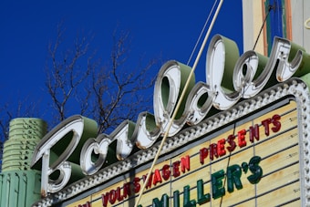 A retro-style marquee sign with the word 'Boulder' in large cursive font, surrounded by LED bulbs. A blue sky and leafless tree branches are visible in the background. The sign also includes words in red and green letters, such as 'VOLKSWAGEN PRESENTS' and 'MILLER'S'. The structure has an Art Deco design, featuring pastel colors like mint green.