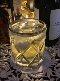 A glass tumbler filled with a clear beverage and ice cubes sits on an intricately patterned tablecloth. The glass has a diamond-cut design, reflecting light in a warm ambient setting. Partially visible liquor bottles surround the glass, creating an elegant and slightly festive atmosphere.