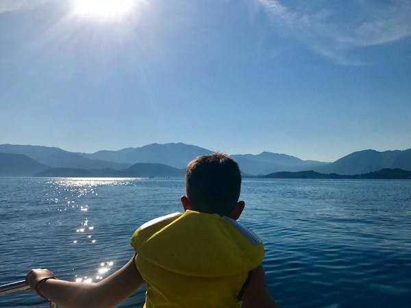 Child in a yellow foam life jacket seen from behind, looking out over sunlit blue water from a boat toward distant mountains