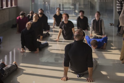 Several people are positioned on a reflective floor in a well-lit room, performing a yoga pose. They are arranged in a spread-out formation, each with a focused and serene expression, looking upward as they stretch their bodies.