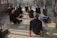 Several people are positioned on a reflective floor in a well-lit room, performing a yoga pose. They are arranged in a spread-out formation, each with a focused and serene expression, looking upward as they stretch their bodies.