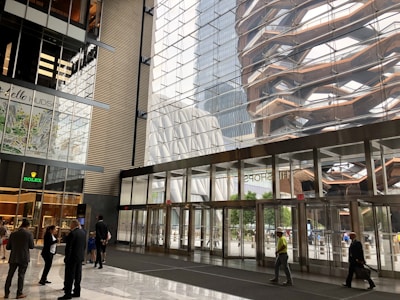 A modern interior space features large glass windows reflecting an intricate outdoor structure. The area has a high ceiling with sleek architectural lines, and several people are walking or standing, suggesting a busy environment. The storefronts include a Rolex sign and other retail displays.