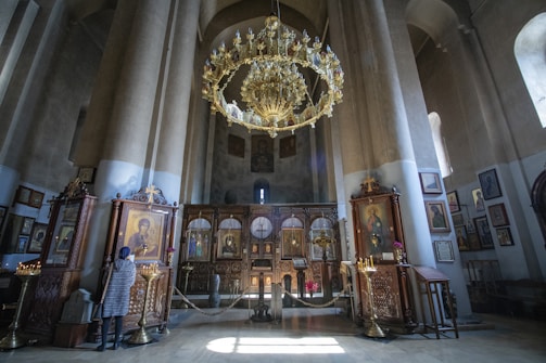 A large, ornate chandelier hangs in a grand room filled with tall pillars and religious icons. The walls are adorned with framed pictures and paintings, and a light softly illuminates the interior. A person stands near a display of candles and religious artwork, dressed in a coat and headscarf. The setting exudes a historical and sacred atmosphere, typical of an Orthodox church.