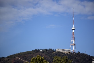 A tall television or radio tower stands prominently on a lush, green hill against a clear blue sky. The structure features a combination of white and red colors, with a circular array around its midsection. Surrounding the tower, there is a building and dense forested area.