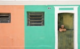 An elderly person with gray hair leans casually over the bottom half of a wooden door. The building wall is split into two colors—peach on the left and teal on the right. Above the door, a small house number plaque is visible. Two windows with horizontal bars are located on the peach and teal sections respectively.
