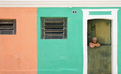 An elderly person with gray hair leans casually over the bottom half of a wooden door. The building wall is split into two colors—peach on the left and teal on the right. Above the door, a small house number plaque is visible. Two windows with horizontal bars are located on the peach and teal sections respectively.