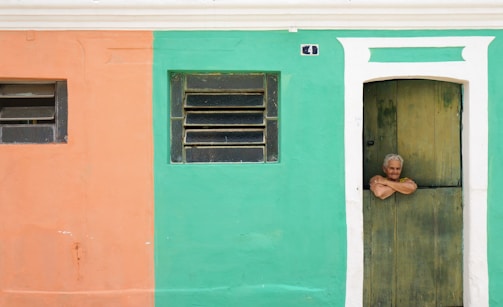 An elderly person with gray hair leans casually over the bottom half of a wooden door. The building wall is split into two colors—peach on the left and teal on the right. Above the door, a small house number plaque is visible. Two windows with horizontal bars are located on the peach and teal sections respectively.