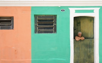 An elderly person with gray hair leans casually over the bottom half of a wooden door. The building wall is split into two colors—peach on the left and teal on the right. Above the door, a small house number plaque is visible. Two windows with horizontal bars are located on the peach and teal sections respectively.