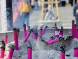 Several bundles of vibrant pink incense sticks are placed upright in a bed of ashes. Smoke is gently rising from the burning tips of the incense, creating a serene and spiritual ambiance. In the background, there are blurred figures which suggest a place of worship or a temple setting.