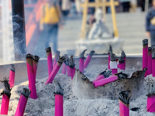 Several bundles of vibrant pink incense sticks are placed upright in a bed of ashes. Smoke is gently rising from the burning tips of the incense, creating a serene and spiritual ambiance. In the background, there are blurred figures which suggest a place of worship or a temple setting.