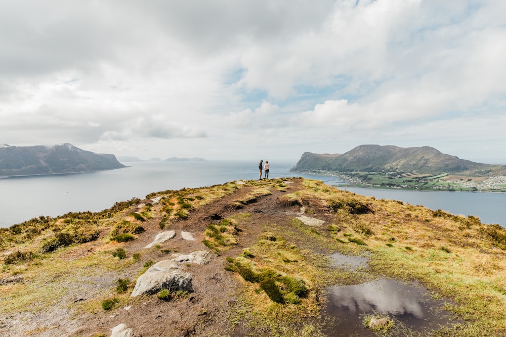 two persons standing near the cliff facing the ocean and mountains during day