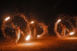 Performers engage in a fire dance, skillfully twirling large fiery torches in circular motions, creating captivating patterns of light against a dark backdrop. The bright flames and flying sparks illuminate their movements as an audience watches the mesmerizing display.