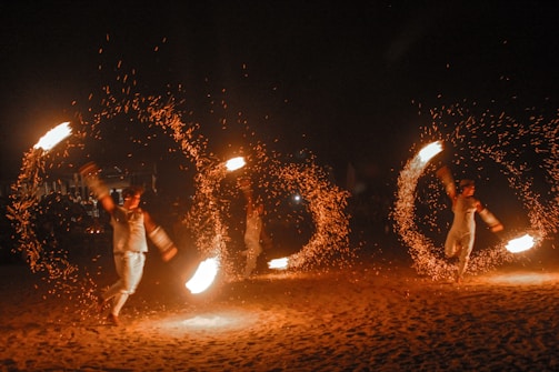 Performers engage in a fire dance, skillfully twirling large fiery torches in circular motions, creating captivating patterns of light against a dark backdrop. The bright flames and flying sparks illuminate their movements as an audience watches the mesmerizing display.