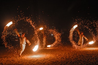 Performers engage in a fire dance, skillfully twirling large fiery torches in circular motions, creating captivating patterns of light against a dark backdrop. The bright flames and flying sparks illuminate their movements as an audience watches the mesmerizing display.